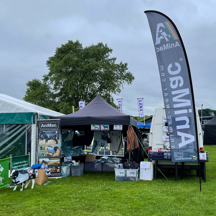 Outdoor trade show booth with a large AniMac flag, banners, and various products displayed under a black canopy tent on grass. There are posters, boxes, and a model animal, with trees and other stalls in the background.