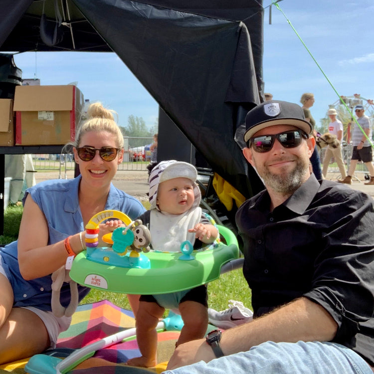 A smiling woman, a baby in a walker, and a man wearing sunglasses and a cap sit together on a blanket outdoors at a sunny event, with tents, boxes, and people visible in the background.