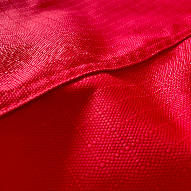Close-up of the textured red fabric from the Airflow Between Combo Waterproof Lightweight Turnout, showing a diagonal seam, a pattern of small dots, a subtle grid, and durable stitching that highlights the material's quality.
