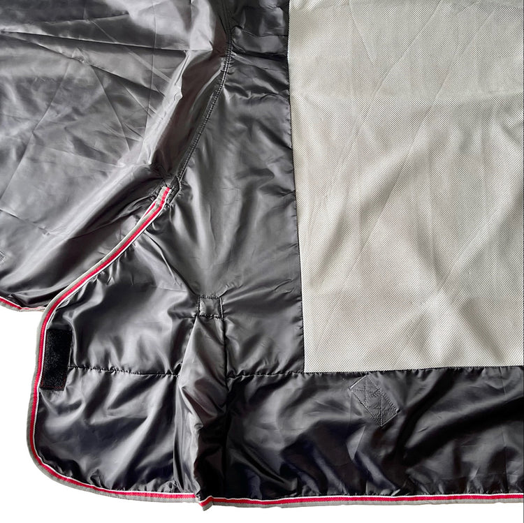 A close-up of the Airflow Between Combo | Waterproof Lightweight Turnout shows black, quilted fabric with a shiny finish, red piping, and light gray mesh for breathability. A small black Velcro strip is visible on the left.