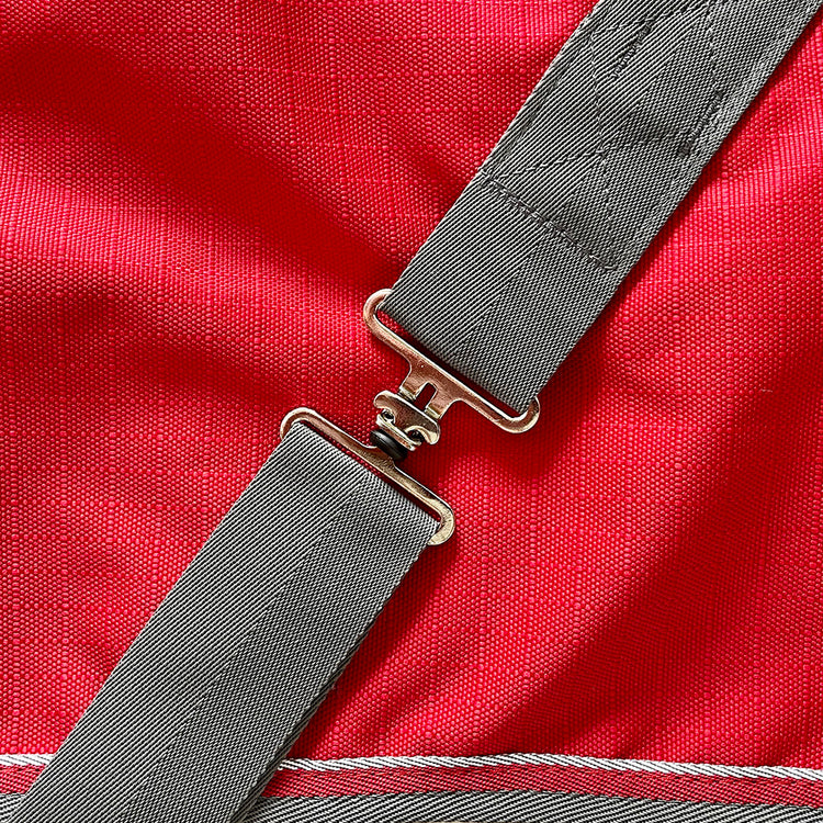 A close-up of the gray adjustable strap with a metal buckle on bright red textured fabric, featured on the Airflow Between Combo Waterproof Lightweight Turnout horse rug.