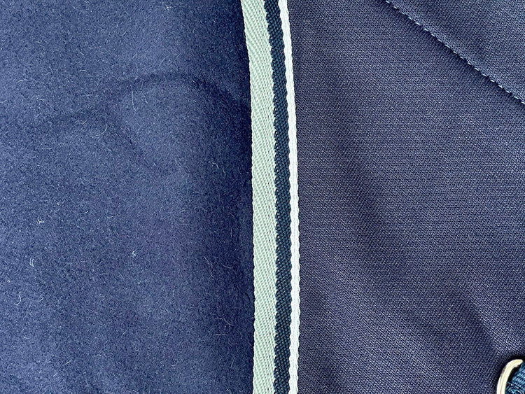 Close-up of two dark blue fabrics from the AniMac Drying Sheep Coat | Wicking/ Cooler, separated by a white and dark green striped trim; a metal button on the right hints at an adjustable fastening feature.