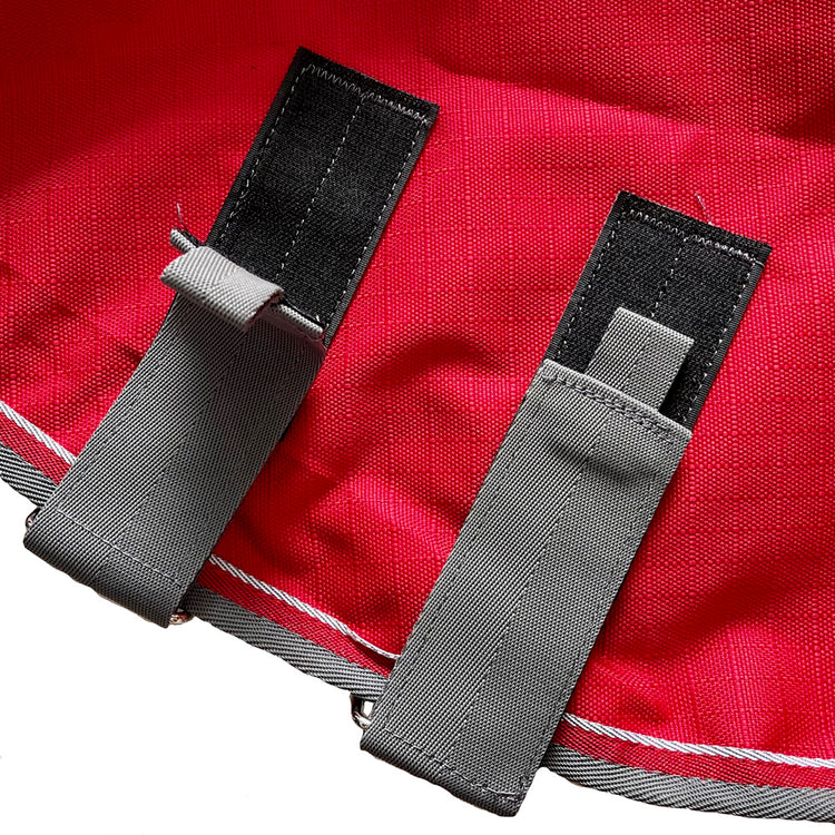 Close-up of two gray fabric straps with black stitched bases on a red surface, resembling fastenings on the Airflow Between Combo | Waterproof Lightweight Turnout rug.